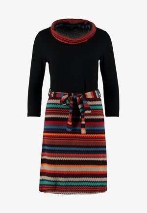 Frau trägt ein schwarzes Top, mehrfarbigen gestreiften Strickrock mit Gürtel, schwarze Strumpfhosen, schwarze Stiefeletten und eine schwarze Umhängetasche mit Kettenriemen.