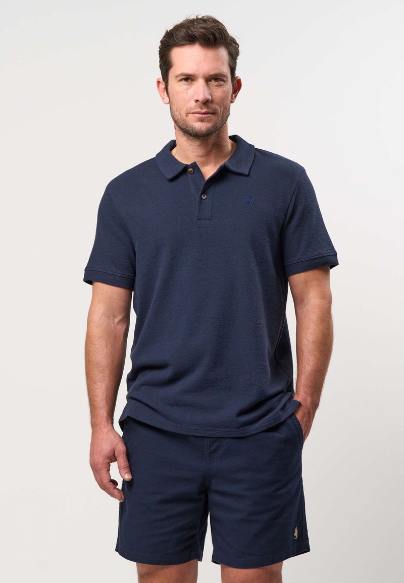 Homme aux cheveux courts et bruns portant un polo bleu marine et un short bleu marine sur un fond clair uni.