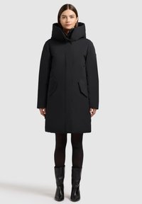 Sort polstret frakke med høj krave, to sidelommer og et strømlinet design, kombineret med knælange sorte støvler. Glat tekstur.