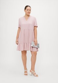 Κοντό ροζ φόρεμα με κοντά μανίκια, V-λαιμόκοψει και φουσκωτή φούστα με επίπεδα. Συνδυασμένο με λευκά σανδάλια με χοντρό τακούνι και ασημένια τσάντα.