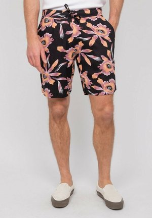 Homme portant un short noir à grands motifs floraux roses et orange, des chaussures blanches sans lacets et une chemise blanche, debout devant un fond uni.