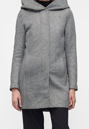 Femme portant un manteau gris à capuche mi-cuisse avec fermeture avant dissimulée et manches longues, associé à un pantalon noir.