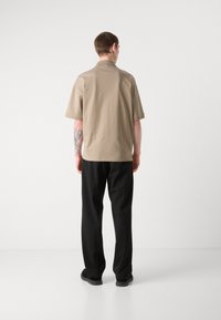 Beige kortermet polo skjorte med krage, kombinert med svarte bukser; glatt stoff og avslappet passform, som viser en tatovering på armen.
