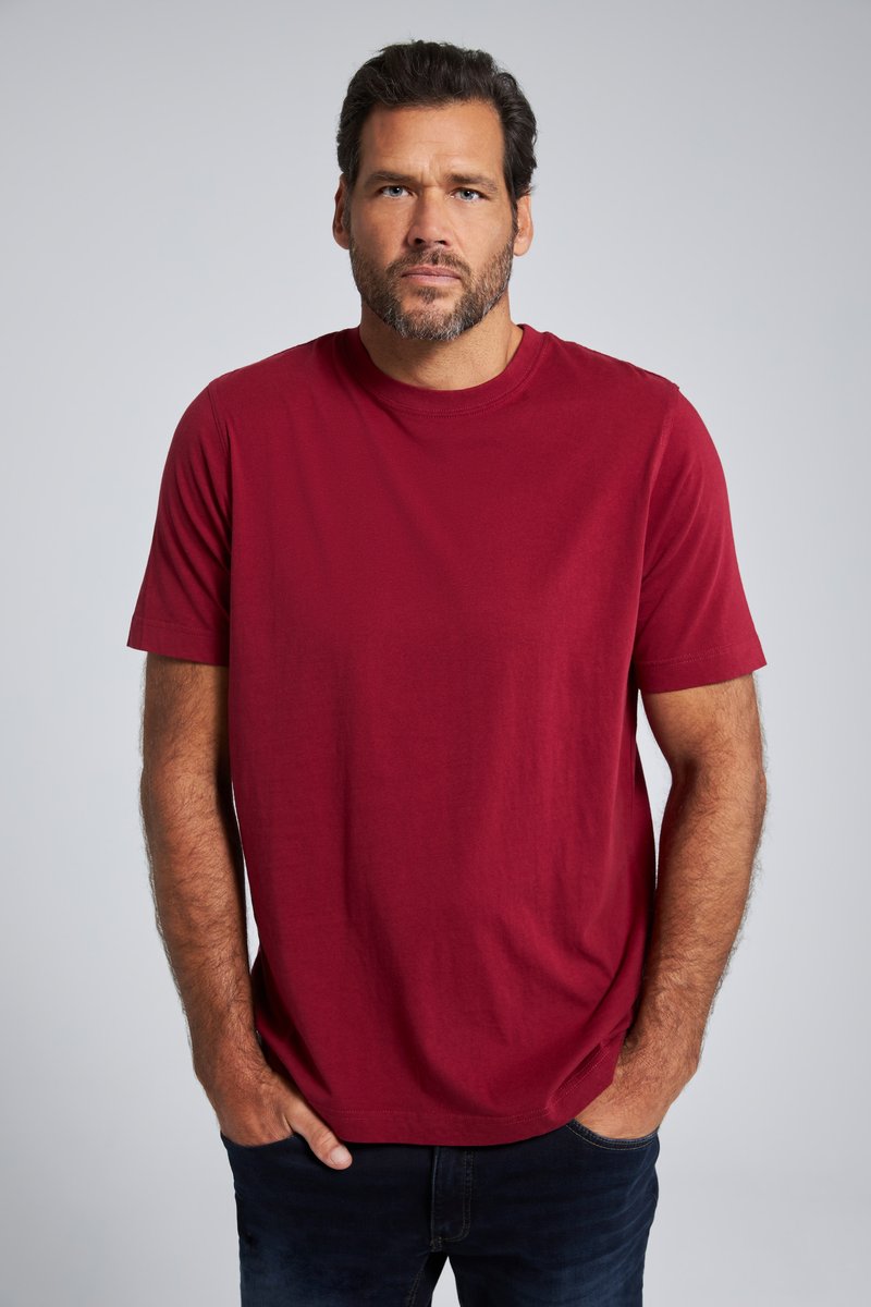 Mann mit Bart und kurzen Haaren, der ein schlichtes rotes T-Shirt und dunkle Hosen trägt, steht mit den Händen in den Taschen vor neutralem Hintergrund.