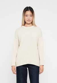 Cremfarbener Strickpullover mit Rundhalsausschnitt, mit überschnittenen Schultern und geripptem Saum. Kombiniert mit dunkelblauen weit geschnittenen Jeans.