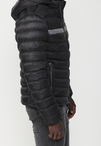 Black puffer jacket featuring horizontal stitching, zippered side pockets, and a hood. Material appears lightweight and water-resistant.