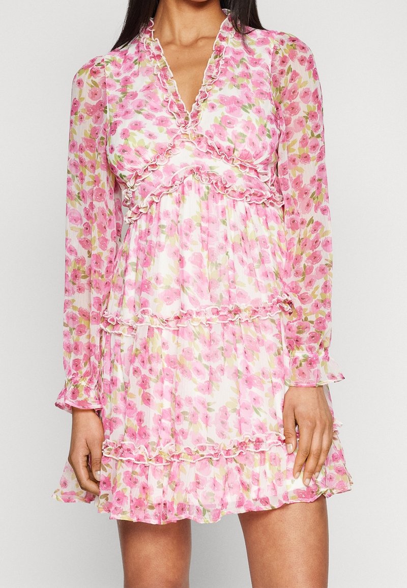 Femme portant une mini-robe à manches longues, imprimé floral rose, avec un col en V à volants et une jupe à volants.