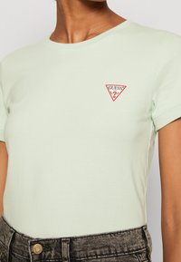 Světle zelené bavlněné tričko s kulatým výstřihem, krátkými rukávy a trojúhelníkovým červeným logem "GUESS?" na hrudi.