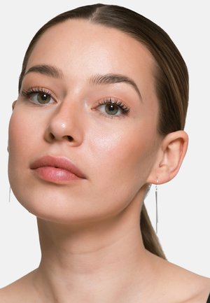 Junge Frau mit heller Haut, zurückgebundenem braunem Haar, trägt dezentes Make-up und silberne Ohrhänger, vor einem einfachen hellen Hintergrund.