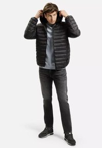 Czarna puchowa kurtka z kapturem, z pikowaną konstrukcją; noszona na jasnoniebieskiej bluzie, zestawiona z ciemnymi dżinsami i trampkami.