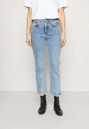 Kvinde iført lyseblå jeans med lige ben, hvid T-shirt og sorte ankelstøvler, stående mod en ensfarvet baggrund.