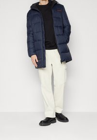 Chaqueta acolchada azul marino con capucha, con secciones acolchadas, cierre con cremallera y un logo rojo. Llevada sobre un suéter negro y combinada con pantalones crema.