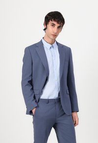 Hellblauer Anzug mit einer taillierten Blazerjacke und Hose, strukturierter Stoff, kombiniert mit einem hellblauen Hemd mit Knopfleiste, schlanke Silhouette.