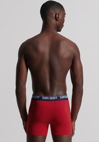 Rode boxershorts met een marineblauwe elastische tailleband waarop 'SUPERDRY' is gedrukt. Soepele textuur, slimfit ontwerp en een middelhoge snit.