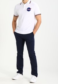 Homme portant un polo blanc de la NASA, un pantalon bleu marine et des baskets blanches, se tenant avec les mains dans les poches devant un fond uni.