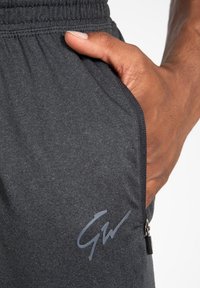 Donkergrijze joggingbroek met een soepele textuur, voorzien van een elastische tailleband, een ritszak en een subtiele GW-logo in het grijs aan de zijkant.