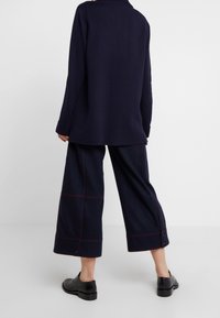 Suéter de gran tamaño de color azul marino con mangas largas y aberturas laterales, combinado con pantalones anchos hasta la rodilla que presentan costuras y botones rojos.
