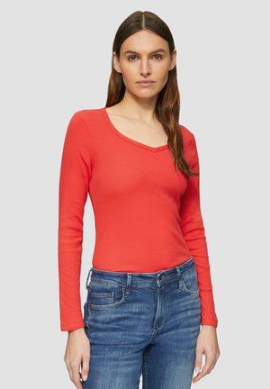 Vrouw met lang bruin haar, gekleed in een rode lange mouw V-hals top en blauwe jeans, staande voor een effen lichtgrijze achtergrond.