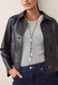 Schwarze Lederjacke mit Kragen, Knopfverschluss und Seitentasche. Darunter ein grauer, gerippter Pullover mit langem Anhänger-Halsband. Dunkle Jeans.