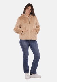 Giacca in pelliccia sintetica beige con cappuccio, chiusura a zip e tasche frontali. La modella indossa jeans blu e sneakers bianche. Tessuto liscio, design accogliente.