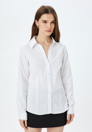 Camicia bianca a maniche lunghe con colletto appuntito, caratterizzata da una trama liscia e una leggera lucentezza, abbinata a una gonna nera.