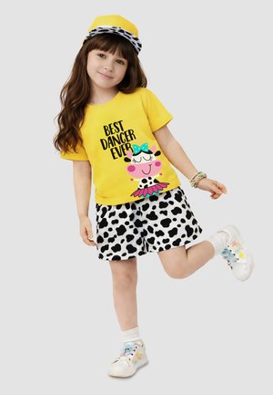 Jeune fille avec un t-shirt jaune « Best Dancer Ever », short imprimé vache, casquette assortie, bracelets colorés, baskets blanches, levant une jambe de manière ludique.