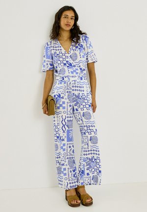 Mujer con un mono de manga corta con estampado azul y blanco, sosteniendo un bolso de mano marrón claro y sandalias de plataforma marrones.