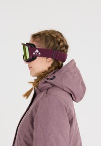 Skibril met een groen gemirroreerde lens, zwarte foam padding en een kastanjebruine band met een witte logo, gedragen met een lichtpaarse jas.