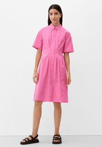Różowa bawełniana sukienka z krótkimi rękawami, zapinana na guziki, z plisowanym fasonem. Połączona z czarnymi sandałami na paski. Prosta i zorganizowana sylwetka.