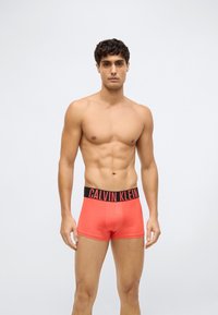 Nuori mies, jolla on lyhyet tummat hiukset, pukeutuneena kirkkaan koralli-huomattaviin bokserimaihinhin, joissa on musta vyötärönauha ja punainen "Calvin Klein" -logo valkoista taustaa vasten.