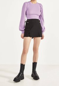 Top lungo a maniche ribbed color lilla con spalle a palloncino, abbinato a una gonna nera plissettata con dettagli in metallo argentato. Stivaletti neri robusti alla caviglia.
