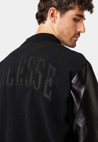 Black bomber jacket featuring wool body and leather sleeves, with large textured lettering on the back and ribbed collar.