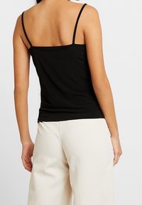 Kvinna som bär en svart ärmlös topp med tunna band och högmidjade vita vida byxor, visar sig från ryggen.