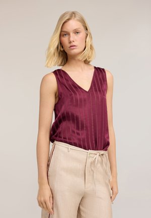 Donna con capelli biondi che indossa un top bordeaux senza maniche con scollatura a V e pantaloni beige a vita alta con fiocco frontale.