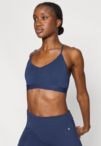 Αθλητικό μπουστάκι σε ναυτικό μπλε χρώμα με σχεδιασμό σε σχήμα V, με ρυτιδωμένη λεπτομέρεια στο μπροστινό μέρος και ρυθμιζόμενες λεπτές τιράντες. Λείο υφή, ελαφρύ ύφασμα.