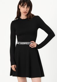 Black long-sleeve dress with a fitted bodice and flared skirt. Features a white branded waistband and sleek, smooth fabric texture.