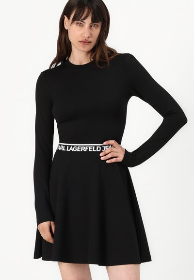 Robe noire à manches longues avec un corsage ajusté et une jupe évasée. Présente une ceinture de marque blanche et une texture de tissu lisse et soyeuse.