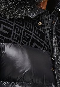 Sort dunjakke med en skinnende, glat overflade og et mønstret fløjlsagtigt øverste lag. Har en sort hætte foret med pels og guldfarvede hardware-detaljer.