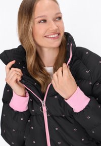 Sort pufferskjorte med pink detaljer, der har lynlås lukning, broderede blomster og ribbede pink manchetter. Hjerteformet lynlåstrækker detalje.