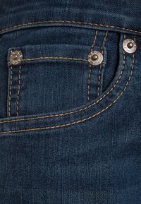 Jeans de mezclilla oscura con un bolsillo frontal que presenta costuras marrones en contraste y dos remaches de bronce, añadiendo durabilidad y detalle.