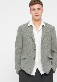 Szary, teksturowany żakiet z wzorem w kratę, dwoma przednimi kieszeniami i klapami. Noszony na białej koszuli, z prostym czarnym guzikiem.