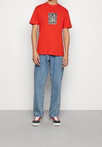 Camiseta de algodón roja con un estampado gráfico de cara en negro y verde en la parte delantera. Pantalones vaqueros de denin azul claro y zapatillas multicolores.