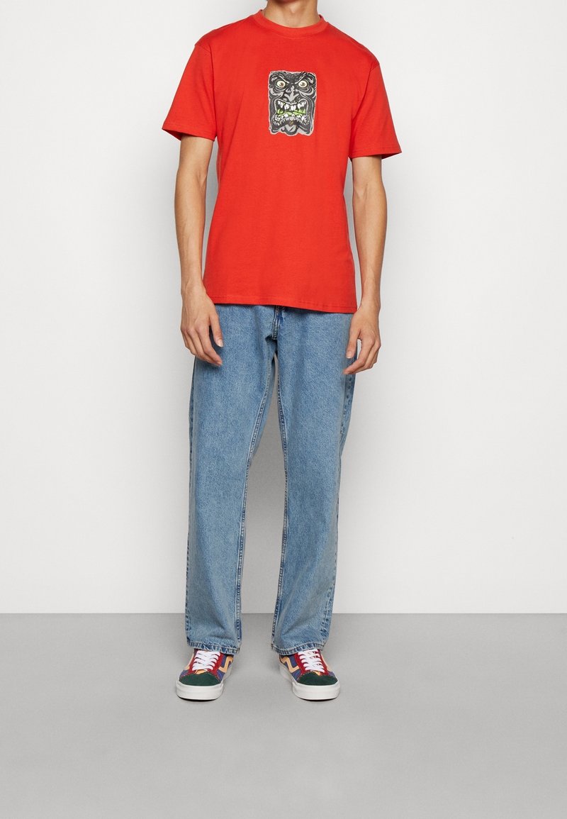 Camiseta de algodón roja con un estampado gráfico de cara en negro y verde en la parte delantera. Pantalones vaqueros de denin azul claro y zapatillas multicolores.