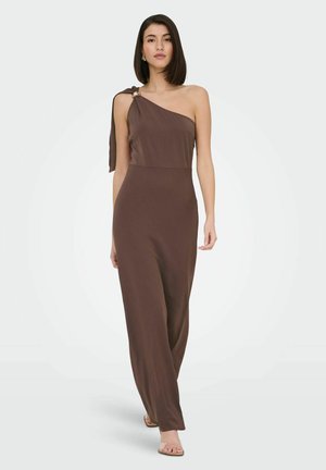 Femme aux cheveux raides et foncés, portant une robe longue marron sans manches à une épaule avec un nœud sur l'épaule, marchant vers l'avant.