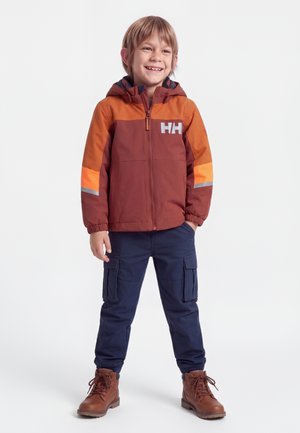Lachende jongen met een bruin-oranje jas, donkerblauwe cargobroek en bruine laarzen, staand met één hand in de zak.