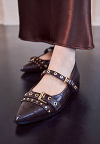 Ballerine marroni con punta affusolata, texture cocco, hardware dorato e cinturini regolabili alla caviglia con accenti circolari.