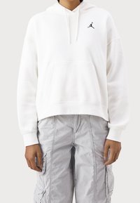 Sudadera corta blanca de tela suave con bolsillo frontal, capucha con cordón y logo negro de Jumpman en el lado izquierdo del pecho. Combinada con pantalones cargo grises.