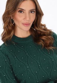 Pulover verde închis cu un model texturat, tricotat în cabluri și perle decorative mici. Guler și mansete cu nervuri. Design elegant și confortabil.