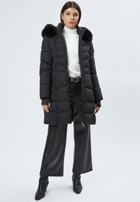 Abrigo acolchado negro con capucha de piel desmontable, con cierre de cremallera y bolsillos laterales, llevado sobre un suéter de punto blanco y pantalones negros.