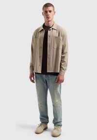 Beige overhemd met knopen en twee voorzakken over een zwart T-shirt, gecombineerd met lichtblauwe distressed jeans en beige schoenen.